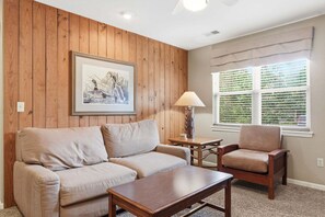 Ein Wohnzimmer mit einer holzverkleideten Wand, einem gerahmten Bild, einem Sofa, einem Sessel, einem Couchtisch und einem Fenster mit Jalousien.