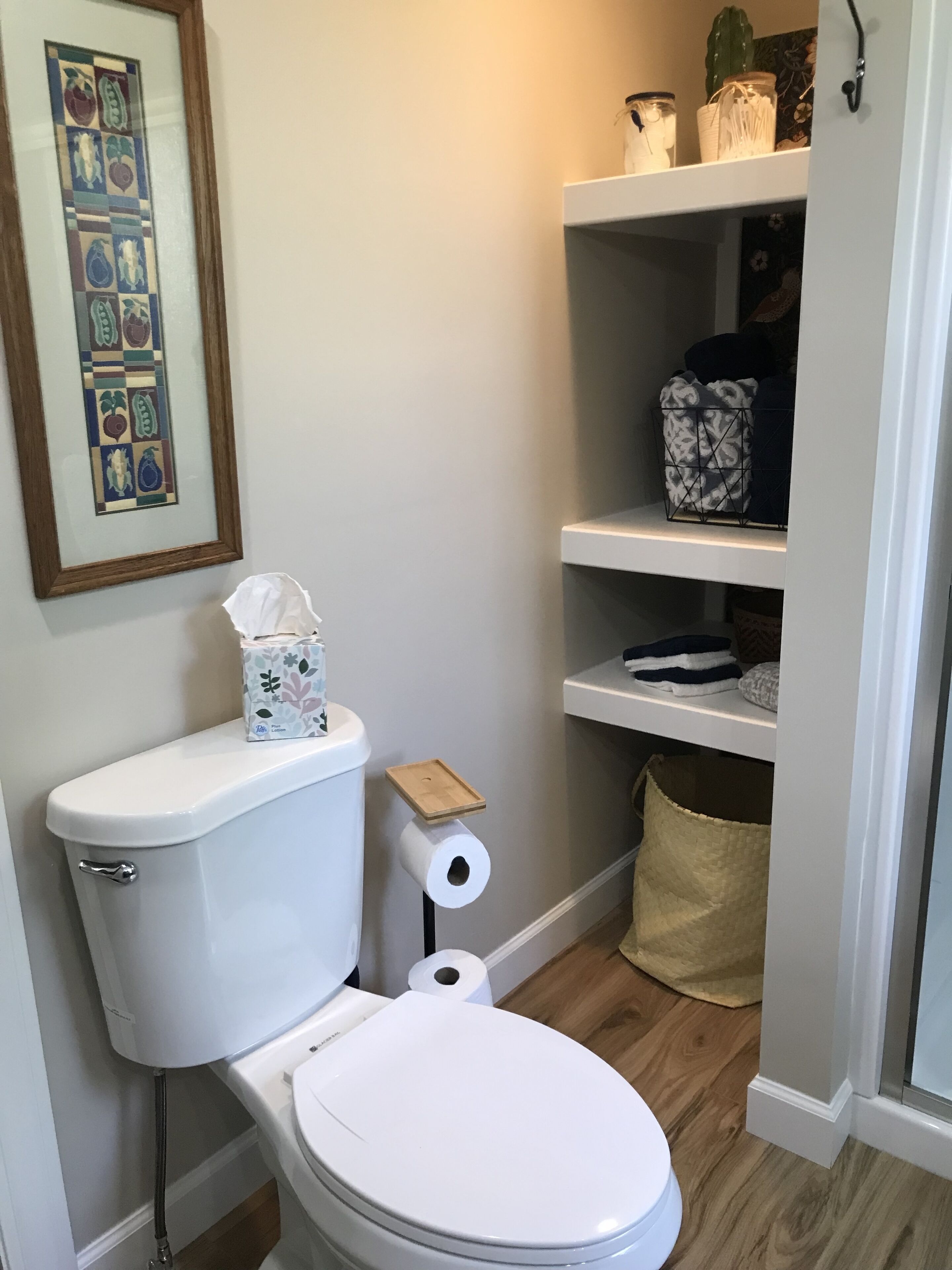 A bathroom with a toilet, a framed artwork on the wall, and a shelf with various items.