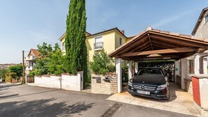 Una calle residencial con un coche estacionado bajo una zona cubierta para vehículos, un alto árbol de hoja perenne y una casa con fachada de piedra y tejado de tejas.