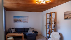 A cozy living room with a wooden ceiling, a dark-colored sofa, a small table, a patterned rug, and a white furry chair. - Vacation home with its own wellness oasis for up to 7 people (Kalübbe)