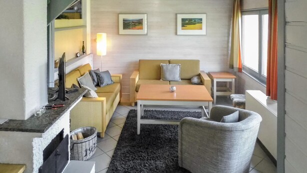 Un salon chaleureux avec un canapé, une table basse et un fauteuil. On y trouve une cheminée, une lampe et deux tableaux encadrés accrochés au mur.