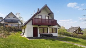 Une maison à deux étages avec un balcon rouge, une antenne parabolique et un petit jardin agrémenté d’un banc.