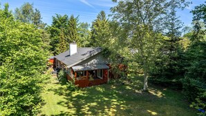 Mökki, jossa on kuisti, ja sen ympärillä on tiheää metsää sekä ruohikkoista pihaa.