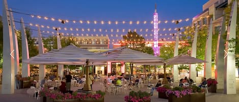 An outdoor dining area with multiple tables and umbrellas, surrounded by flower planters and string lights.