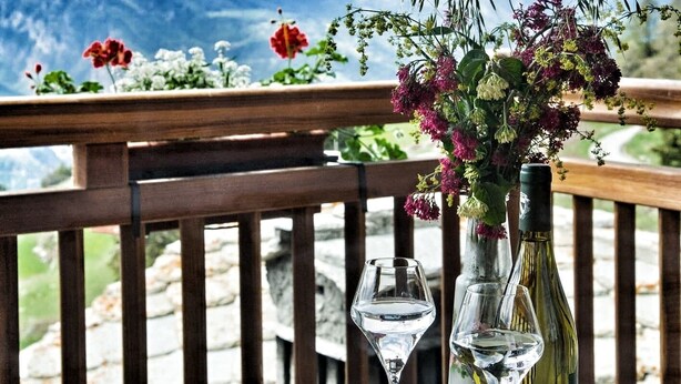 Une table en bois avec un verre et une bouteille de vin, un vase rempli de fleurs et une vue sur des montagnes enneigées.