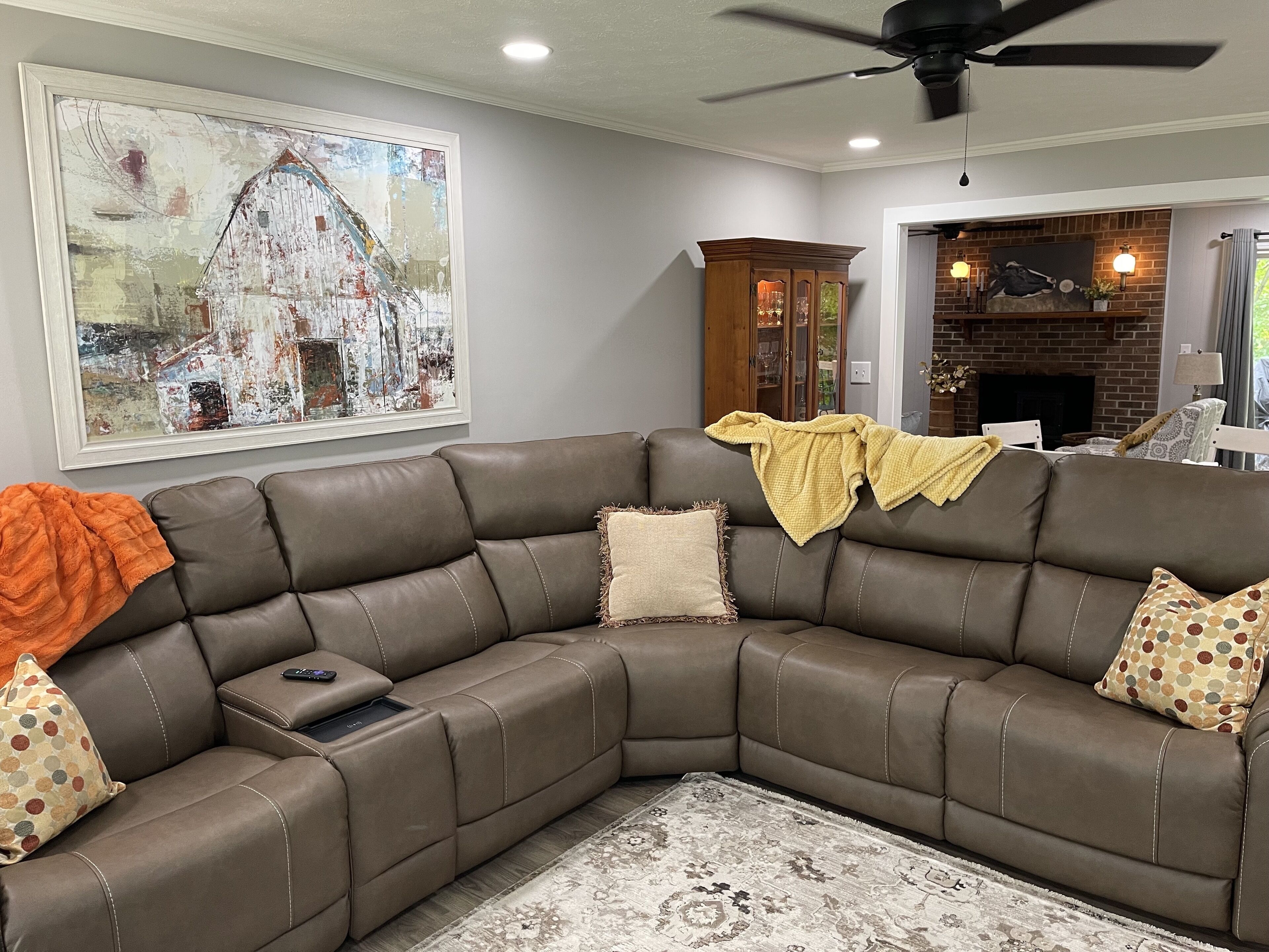 A living room with a brown leather sectional sofa, a large abstract painting, a wooden cabinet, and a brick fireplace.