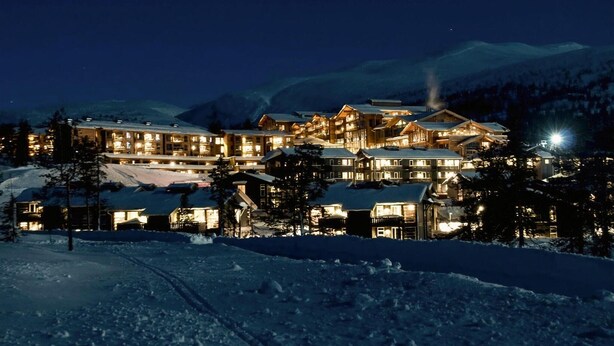 Un chalet enneigé composé de plusieurs bâtiments, avec des fenêtres illuminées et un paysage montagneux en arrière-plan.