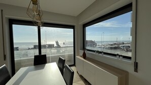 Zona de menjador moderna amb una taula gran i caires, finestres grans amb vista a la platja i l'oceà, i una làmpada penjant.