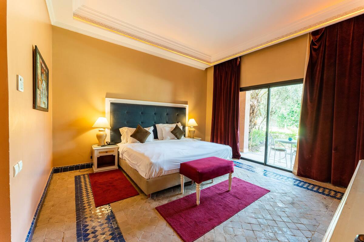 A hotel room with a large bed, two bedside lamps, a red bench, a patterned rug, and a view of greenery through a large window.