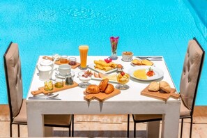 Une table de petit-déjeuner installée au bord d’une piscine, avec divers aliments, boissons et un vase rempli de fleurs.