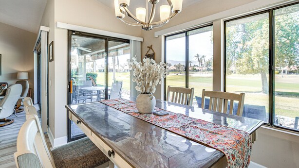 A dining area with a large table, chairs, a vase with flowers, and large windows offering a view of the outdoors.