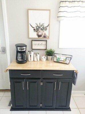 A kitchen counter with a coffee maker, two thermoses, a small potted plant, and a basket of items. Below, there are two black cabinets with handles. - Dog Friendly|Fenced|Winter Monthly Rates (Alpena)