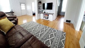 A modern living room with a brown leather sofa, a patterned rug, a flat-screen TV, and a white brick wall.