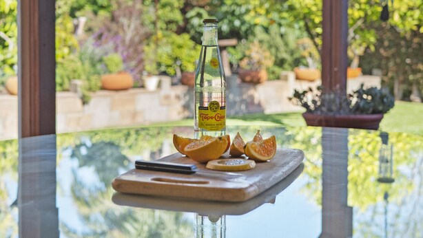 Eine Flasche Mineralwasser Topo Chico auf einem Glastisch zusammen mit einem Holztisch und Orangenscheiben.