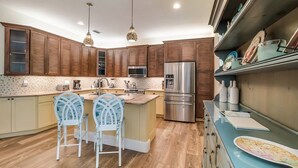 A modern kitchen with wooden cabinets, a stainless steel refrigerator, two bar stools, and pendant lighting. - Pineapple Cay: 2.5 Blocks to Beach! Pool & Spa by Anna Maria Life Vacation Rentals (Holmes Beach)