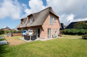 A brick house with a thatched roof, a patio with outdoor furniture, and a well-maintained lawn. - 38/13 Senskiin Lee - Moderne & Nachhaltige Haushälfte Unter Reet mit ca. 110 m² Wohnfläche für 6 (Wenningstedt-Braderup (Sylt))