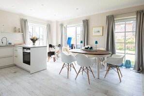 A modern kitchen with white cabinets, a built-in oven, and a dining area with a round table and white chairs. - 38/13 Senskiin Lee - Moderne & Nachhaltige Haushälfte Unter Reet mit ca. 110 m² Wohnfläche für 6 (Wenningstedt-Braderup (Sylt))