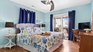 Un dormitorio con cama, dos mesitas de noche, ventilador de techo, televisor y vistas al exterior.