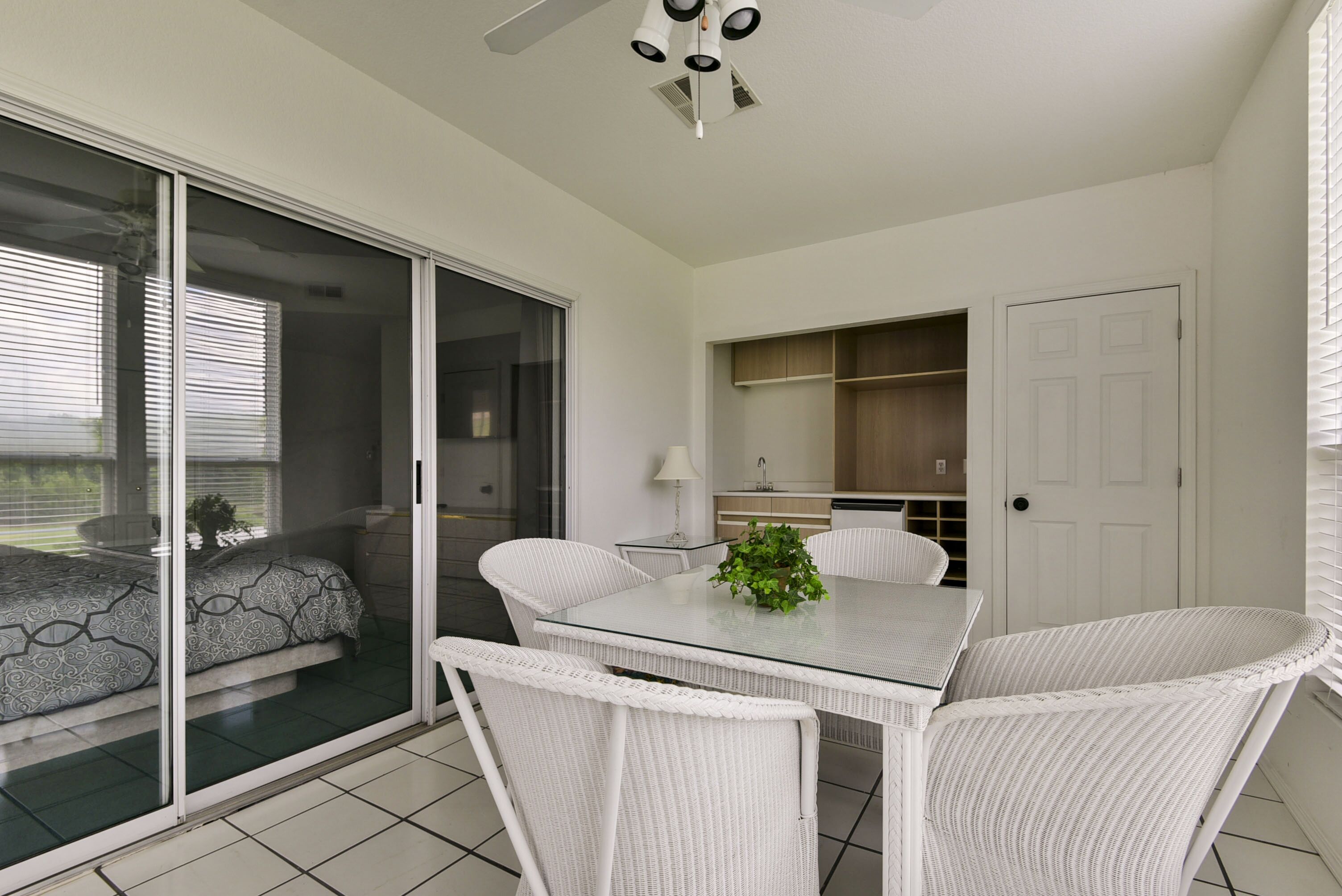 A modern kitchen with white cabinets, a dining area with a glass table and wicker chairs, and a sliding glass door leading to a balcony.