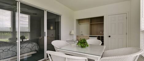 A modern kitchen with white cabinets, a dining area with a glass table and wicker chairs, and a sliding glass door leading to a balcony.