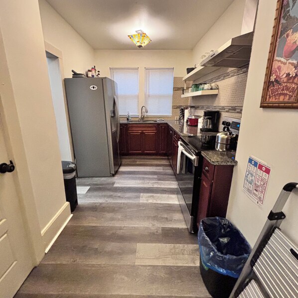 A narrow kitchen with a refrigerator, stove, and sink. There is a trash can and a blue bag on the floor.