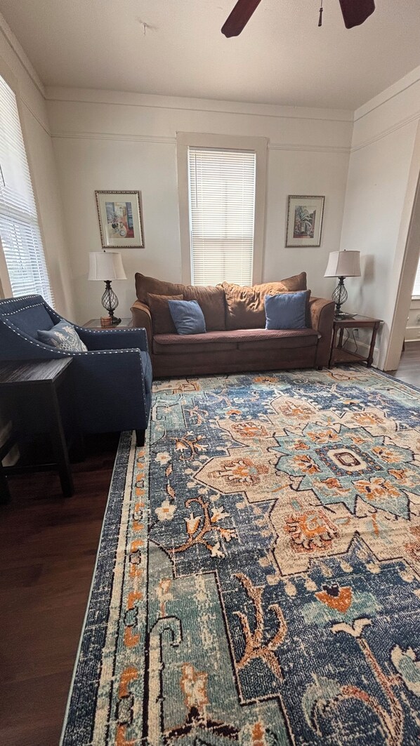 A living room with a sofa, a blue armchair, a patterned rug, a ceiling fan, and two wall-mounted lamps.