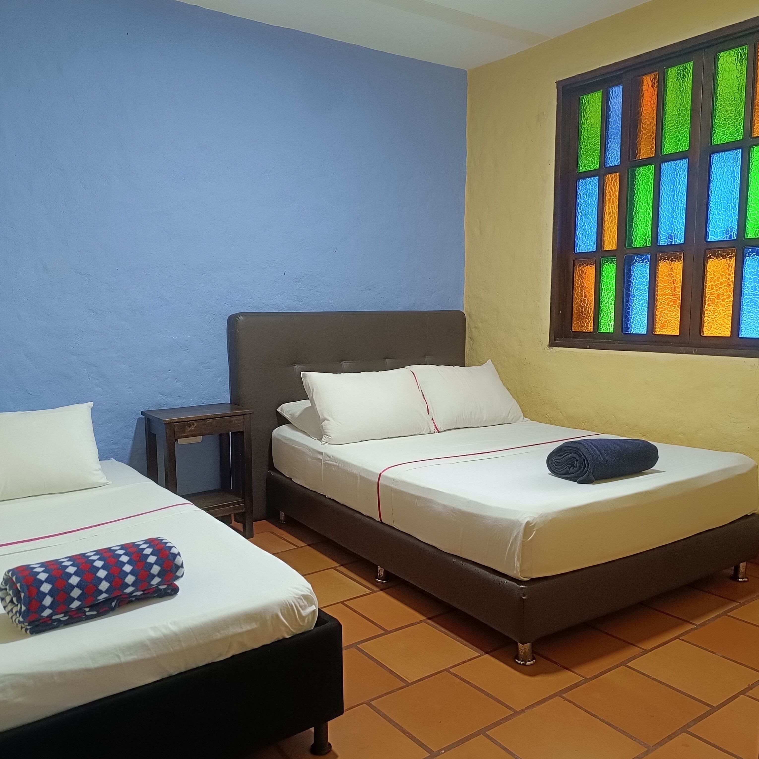 Une chambre avec deux lits, une petite table et une fenêtre ornée de vitraux colorés.