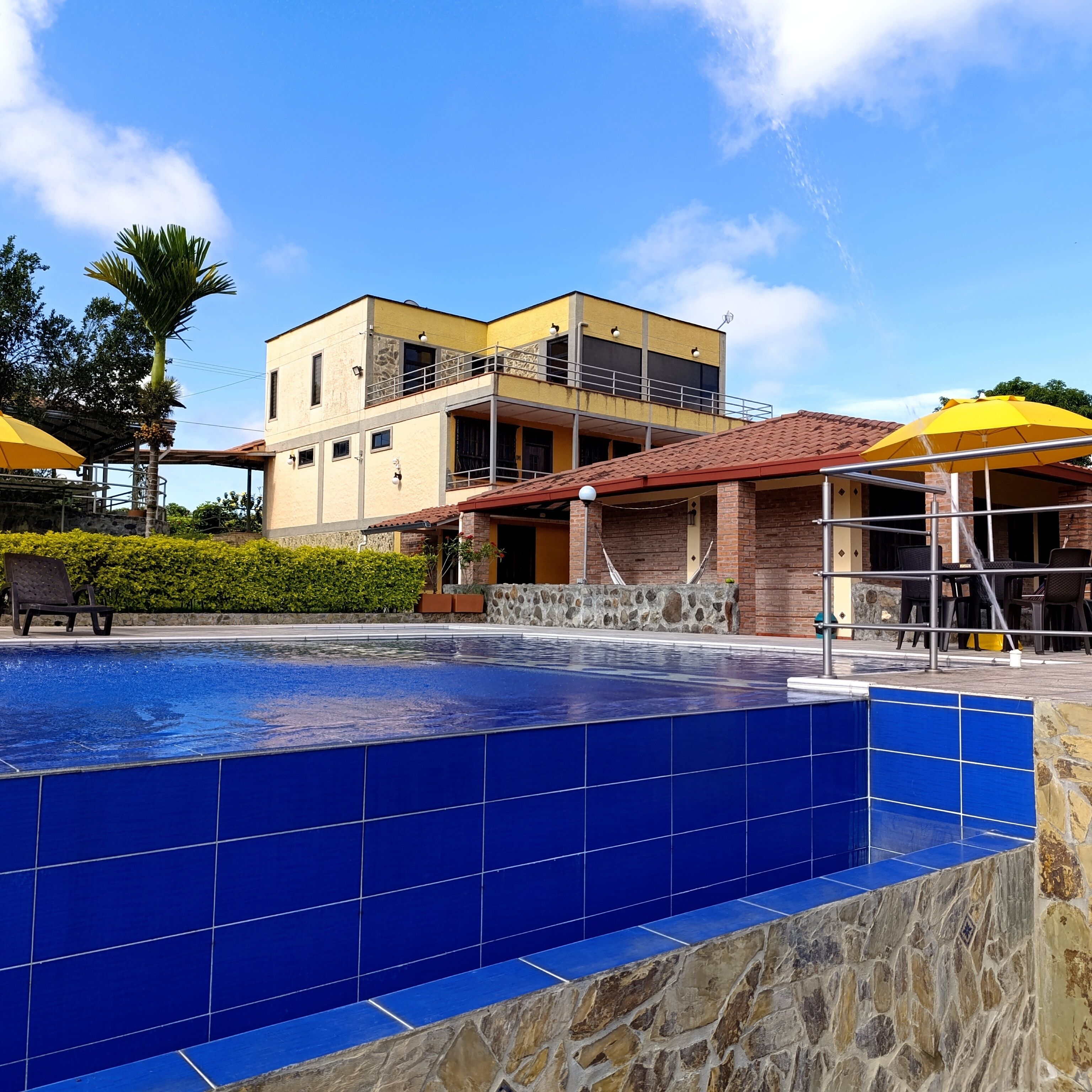 A swimming pool with blue tiles, a yellow building with a balcony, and a yellow umbrella.