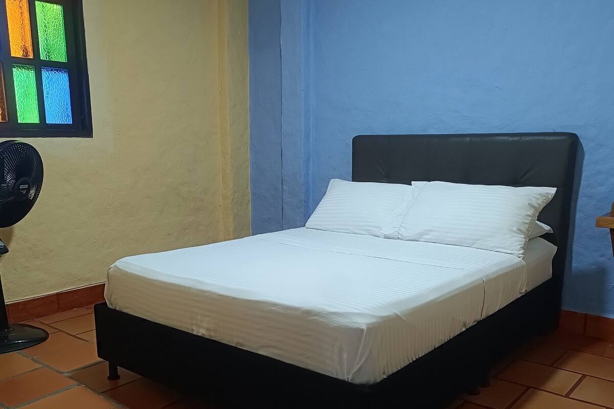 A bed with white bedding in a room with blue walls and a window with colorful glass panes.