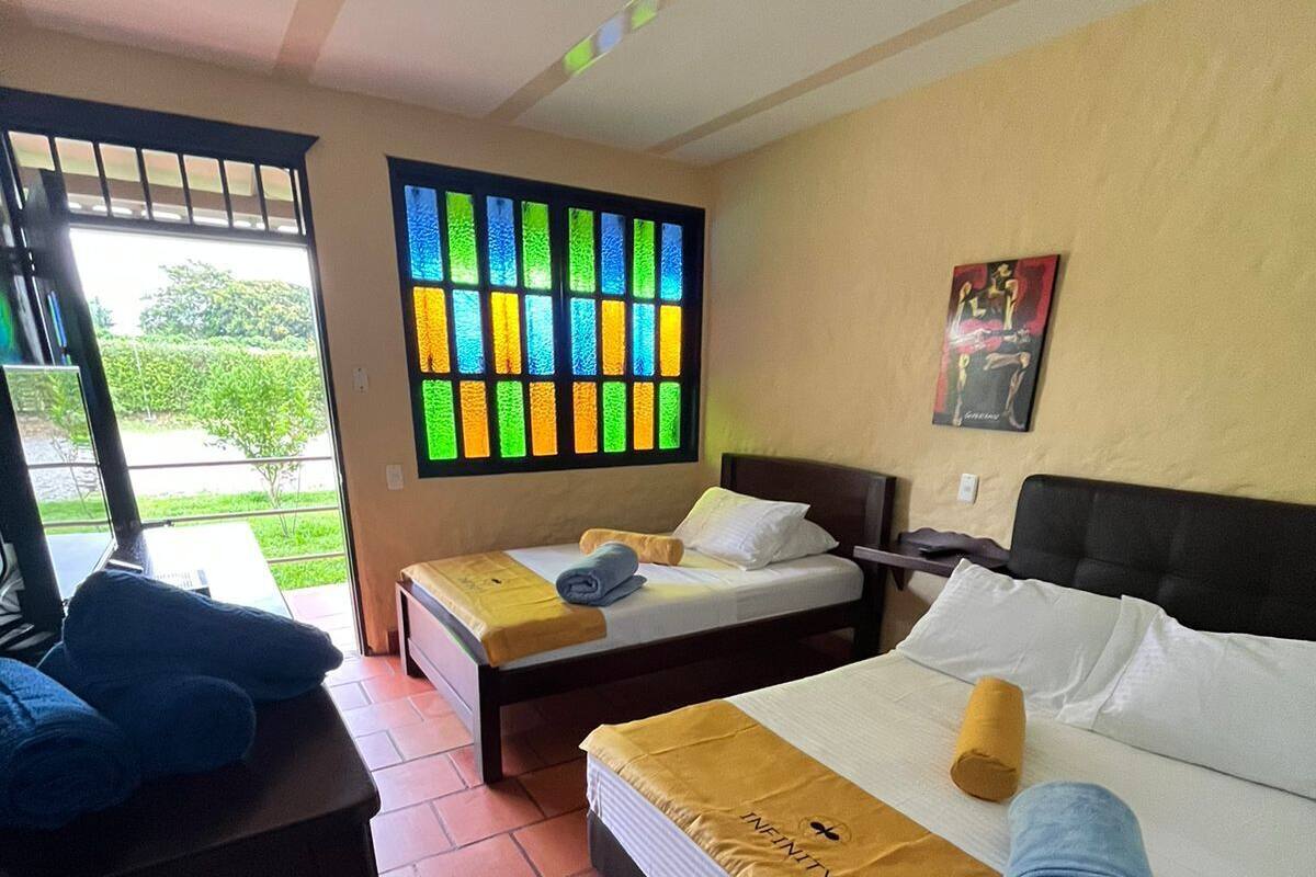 A hotel room with two beds, a window with colorful stained glass, a painting on the wall, and a view of greenery outside.