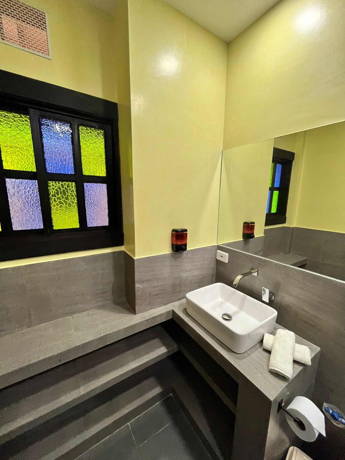 A modern bathroom with a large mirror, a white sink, and a window with colorful stained glass.