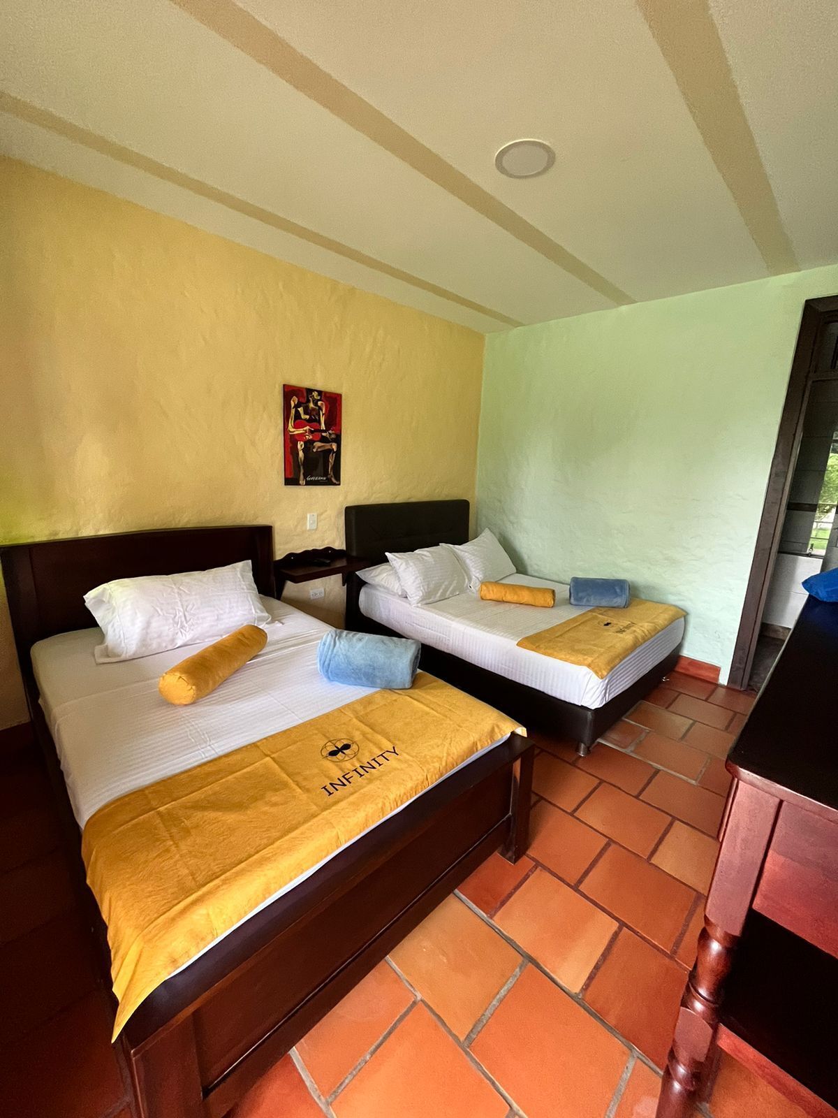Habitación de hotel con dos camas, cada una con una manta y almohadas amarillas, y un pequeño cuadro en la pared.