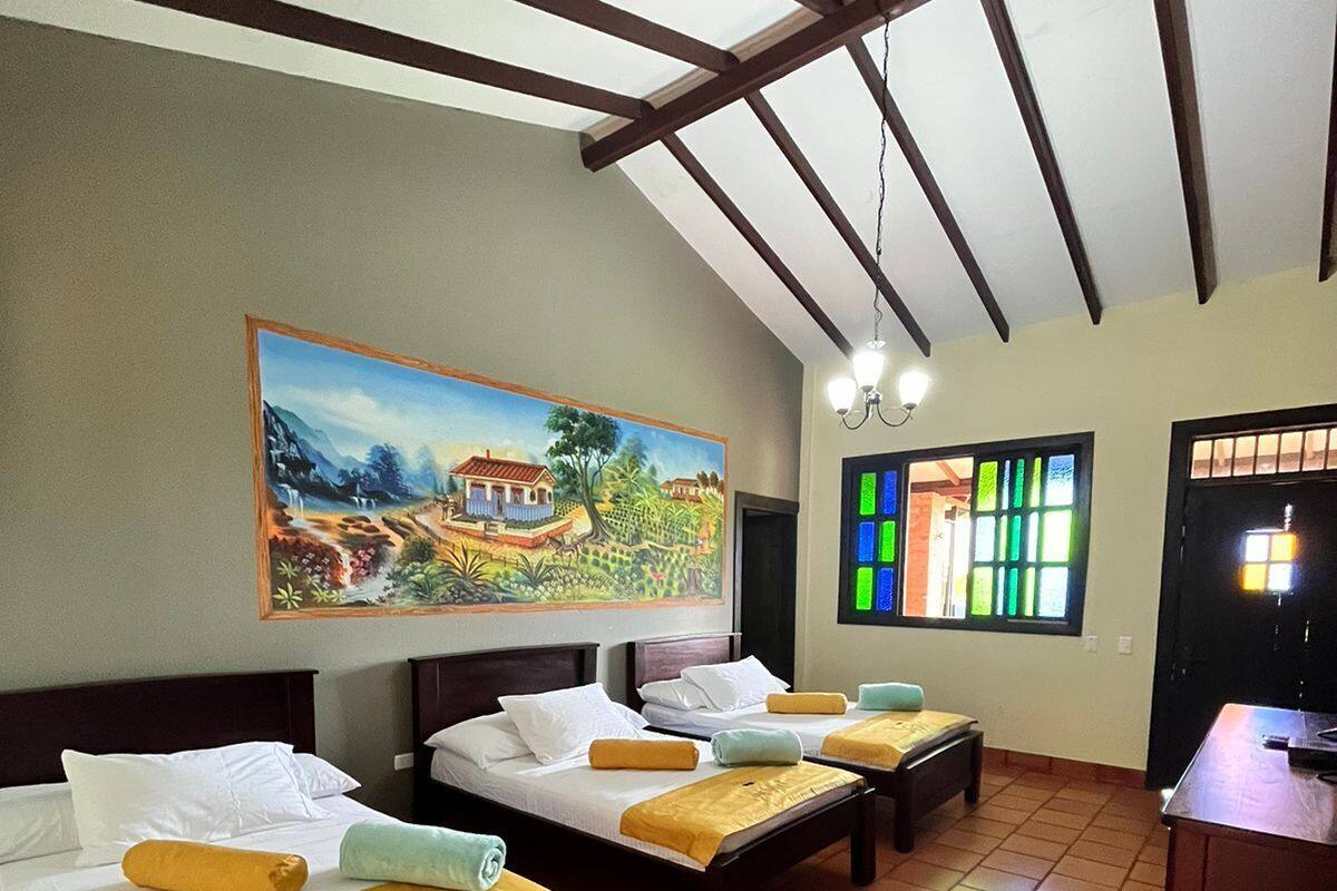 A hotel room with three beds, a painting on the wall, a window with colorful panes, and a tiled floor.