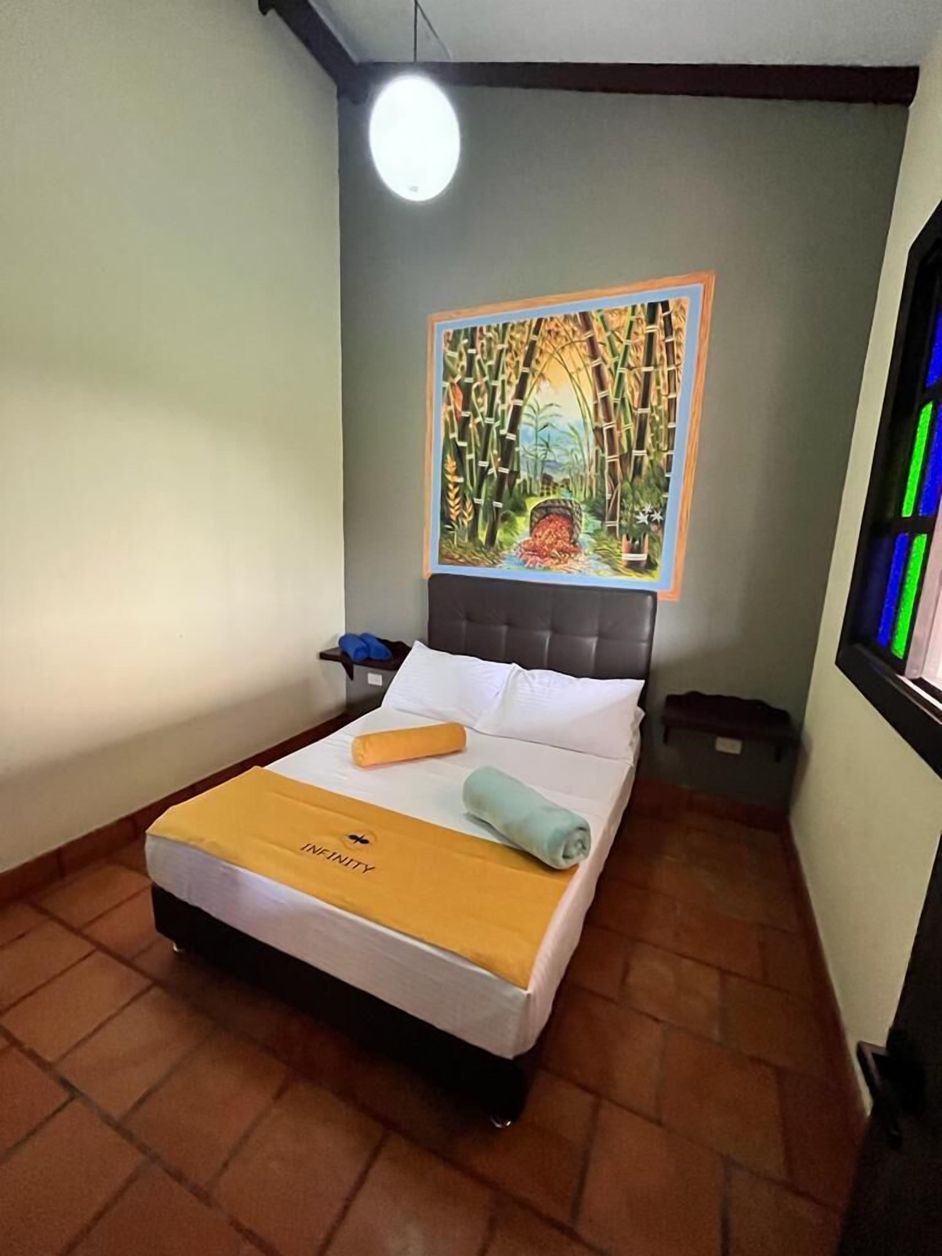 Una habitación de hotel con cama, un cuadro en la pared, una mesita de noche y una ventana con luces de colores.
