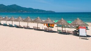 A beach with rows of thatched umbrellas and lounge chairs, a signboard, and a clear view of the sea and distant hills. - Sea Sand 2 Hotel (Da Nang)