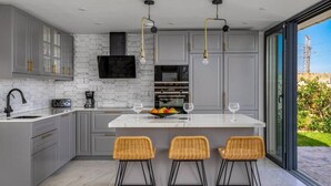 A modern kitchen with grey cabinets, a white island with bar stools, a large window, and a view of greenery outside. - Modern Elegance/Private Pool (Fuengirola)