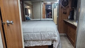A compact bedroom with a bed, wooden flooring, a small kitchenette, and a door leading to another room.