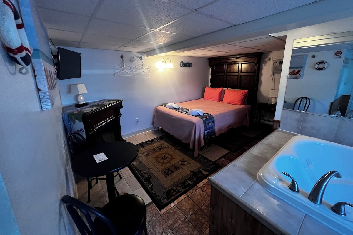 A compact hotel room with a bed, a desk, a chair, a small table, a bathroom with a sink and a bathtub, and a mirror.