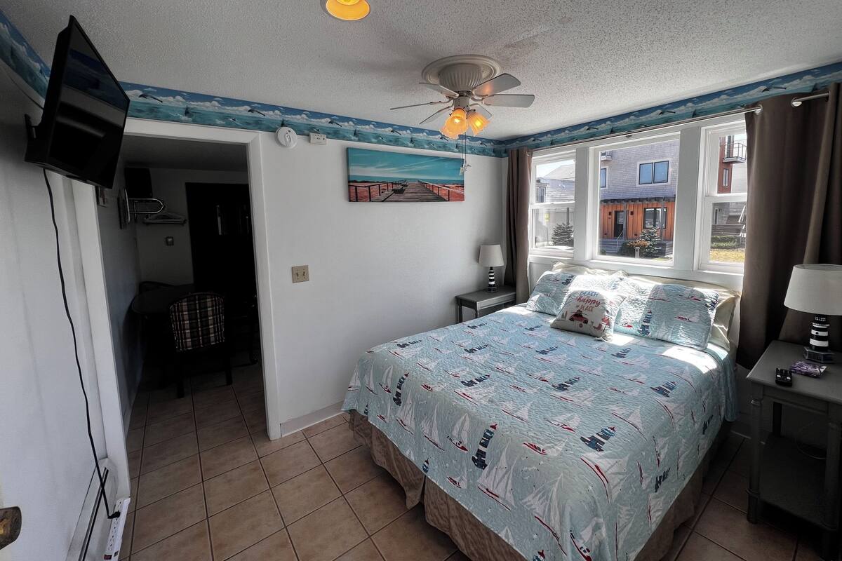 A bedroom with a bed, a ceiling fan, a TV, a window with curtains, and a painting on the wall.
