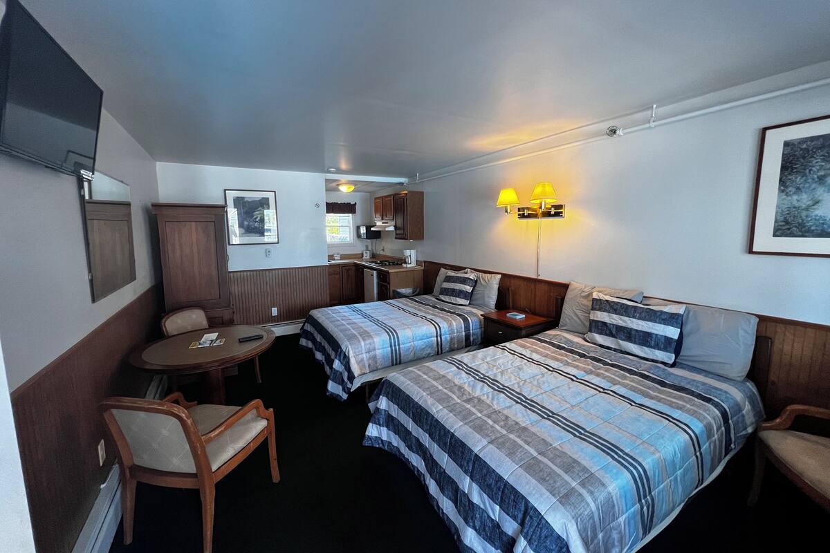 A compact hotel room with two beds, a small kitchenette, a dining table, and a single chair.