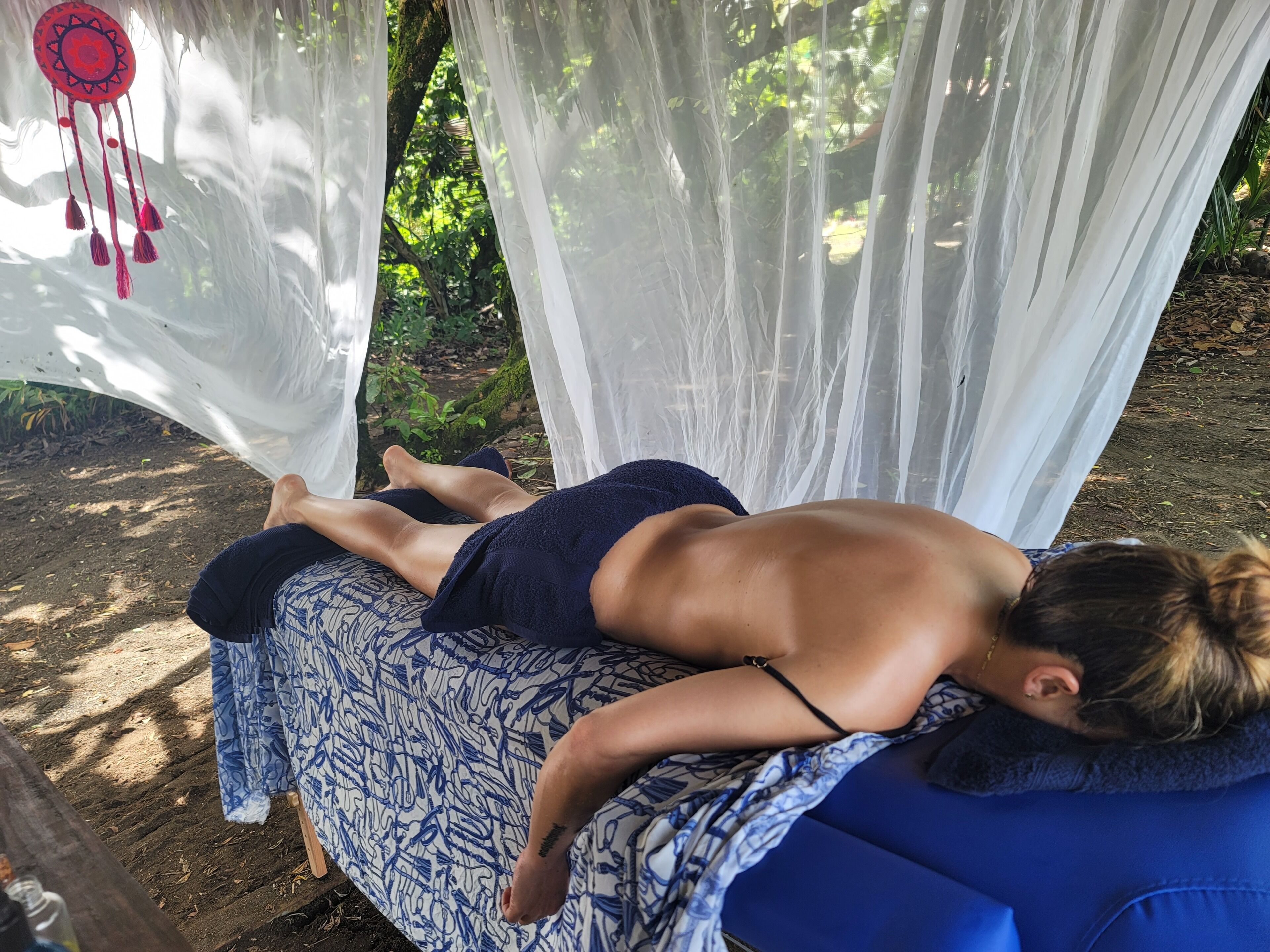A person lying on a blue couch with a patterned blanket, under a canopy with white curtains and a dreamcatcher hanging above.