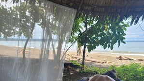 Una persona recostada sobre un colchón estampado bajo un techo de paja, con una playa y palmeras al fondo.