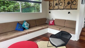 A modern living room with a large window, a woven ceiling light, a woven rug, a red ottoman, a black wicker chair, and a brown sectional sofa with patterned cushions. - Just ONE Minute TO Medlands Beach (Auckland)