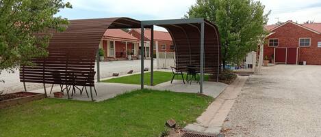 A covered outdoor seating area with benches and a shelter, a gravel driveway, and a brick building in the background.