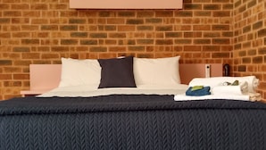 Una cama con una colcha acolchada oscura, dos almohadas y una toalla doblada en una mesita de noche. El fondo de la habitación es una pared de ladrillo.