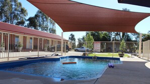Una piscina con zona sombreada, rodeada por una cerca, y un edificio de techo rojo al fondo.