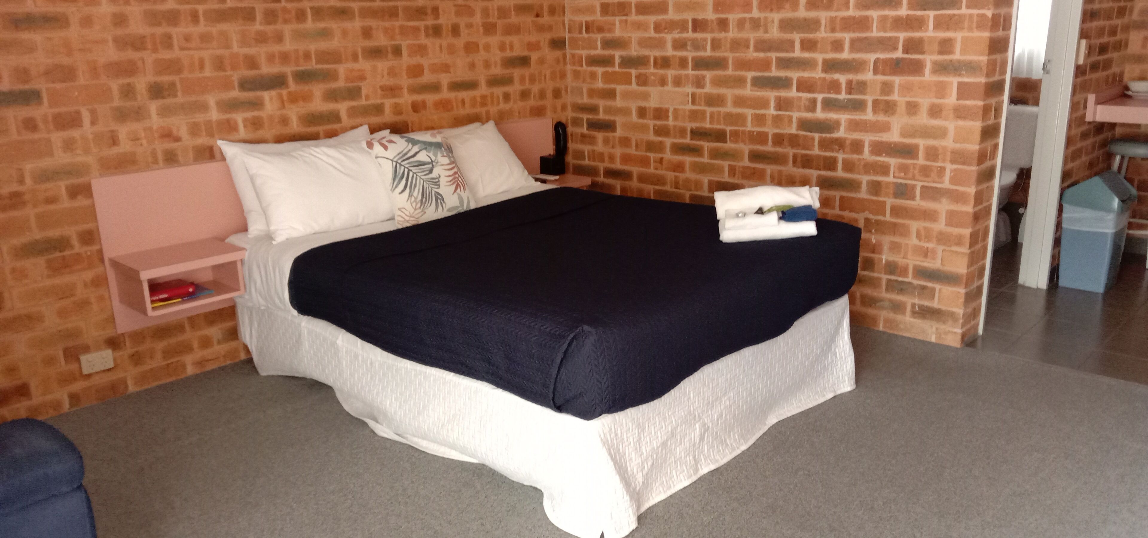Un dormitorio con pared de ladrillo, cama con ropa de cama blanca y azul marino, mesita de noche y estantería pequeña con libros.