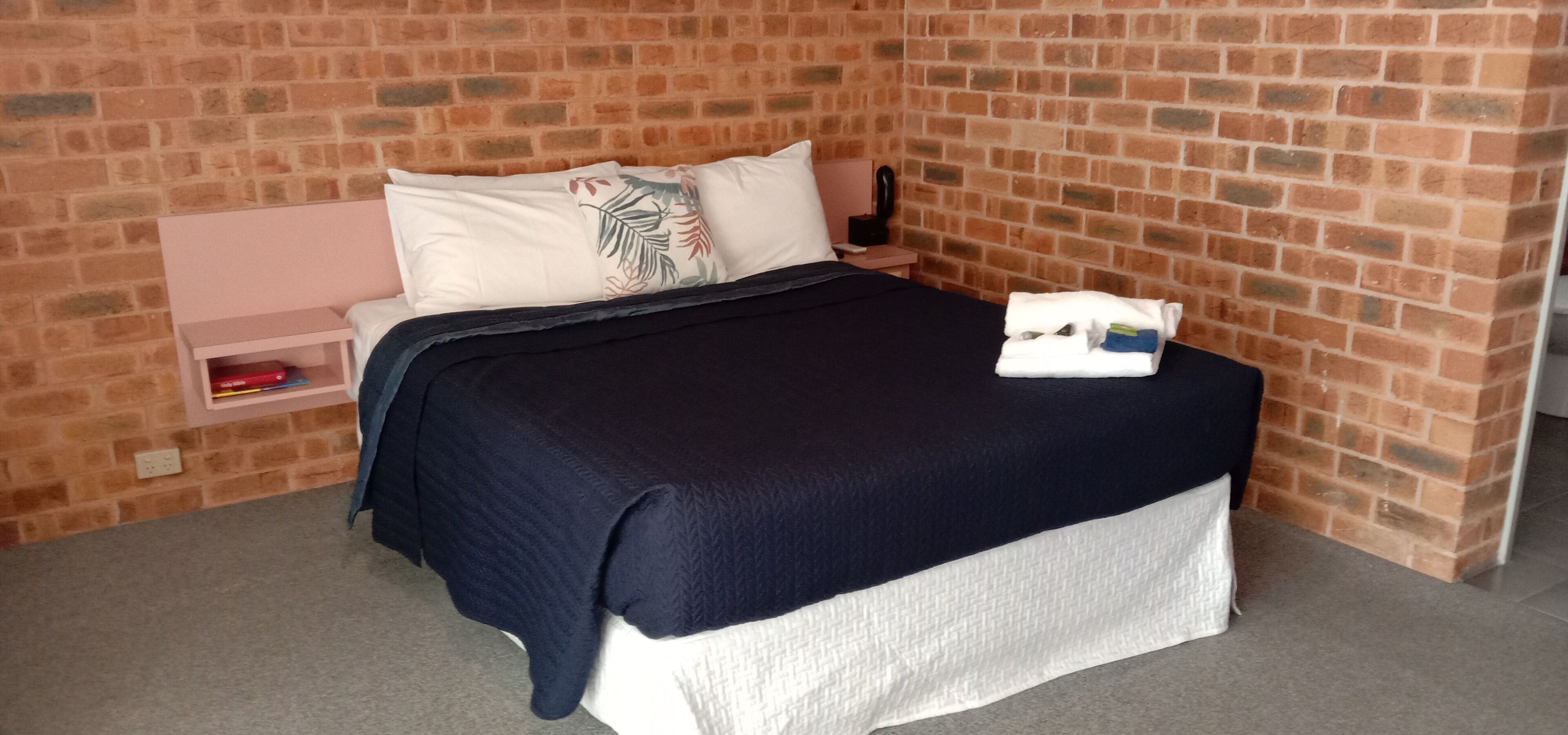 Una cama individual con una colcha azul oscuro y almohadas blancas, una mesita de noche rosa con un teléfono y un libro rojo, y una pared con diseño de ladrillo.