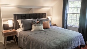 A neatly made bed with a grey quilt, two bedside tables with lamps, a window with curtains, and a red rug. - J Birds' Cottage in the heart of Midtown  (Memphis)