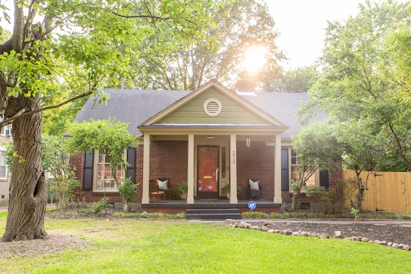 A single-story house with a front porch, a brick facade, and a front yard with a tree and a wooden fence.